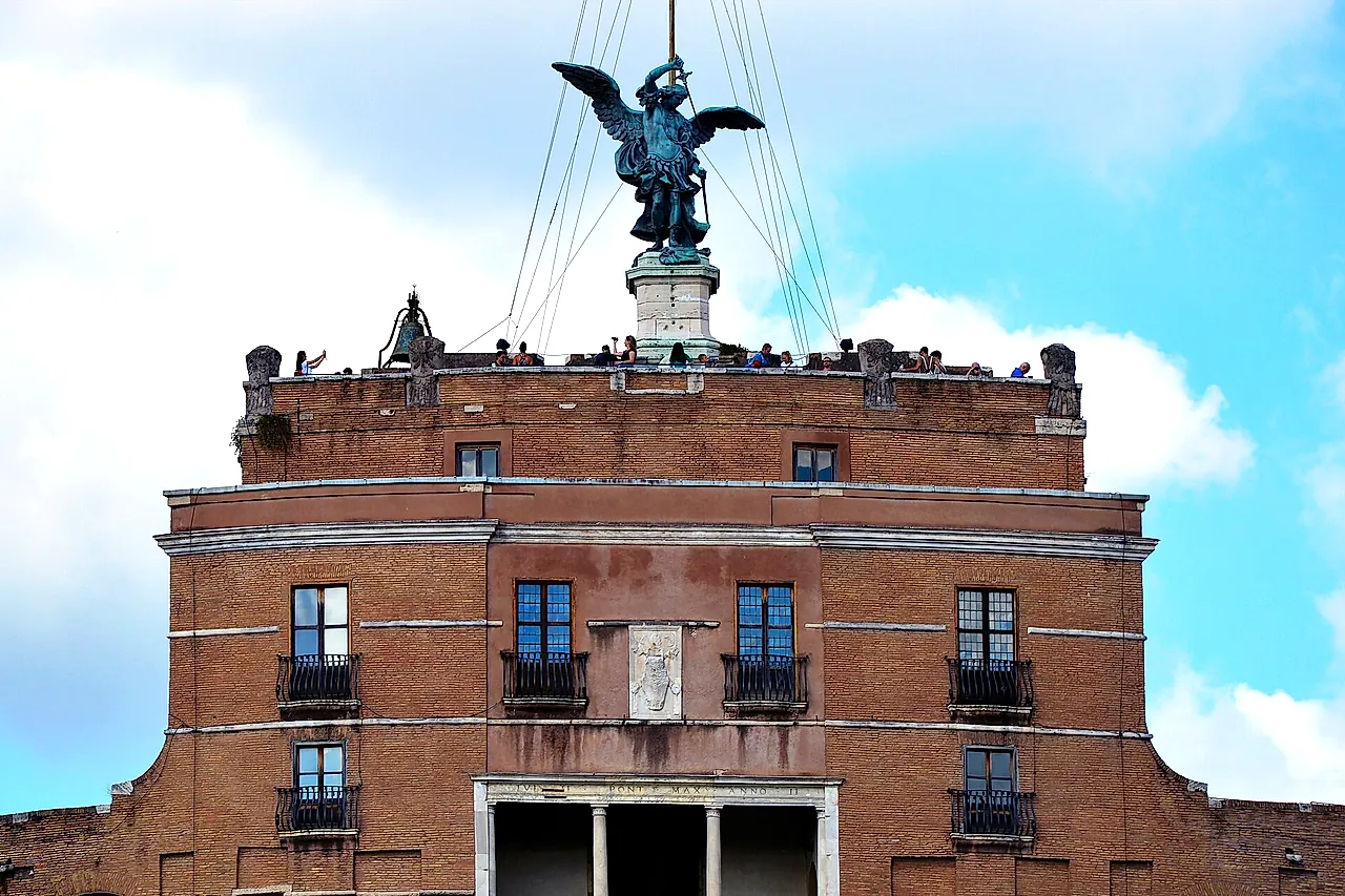 The top of Castel Sant'Angelo: the bronze statue of the Archangel Michael with spread wings on the summit, above the brick upper block with its loggia and the terrace where tiny figures of visitors stand