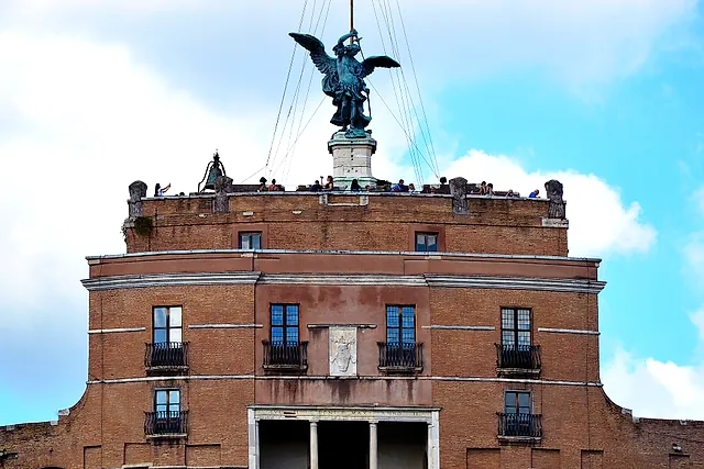 The top of Castel Sant'Angelo: the bronze statue of the Archangel Michael with spread wings on the summit, above the brick upper block with its loggia and the terrace where tiny figures of visitors stand