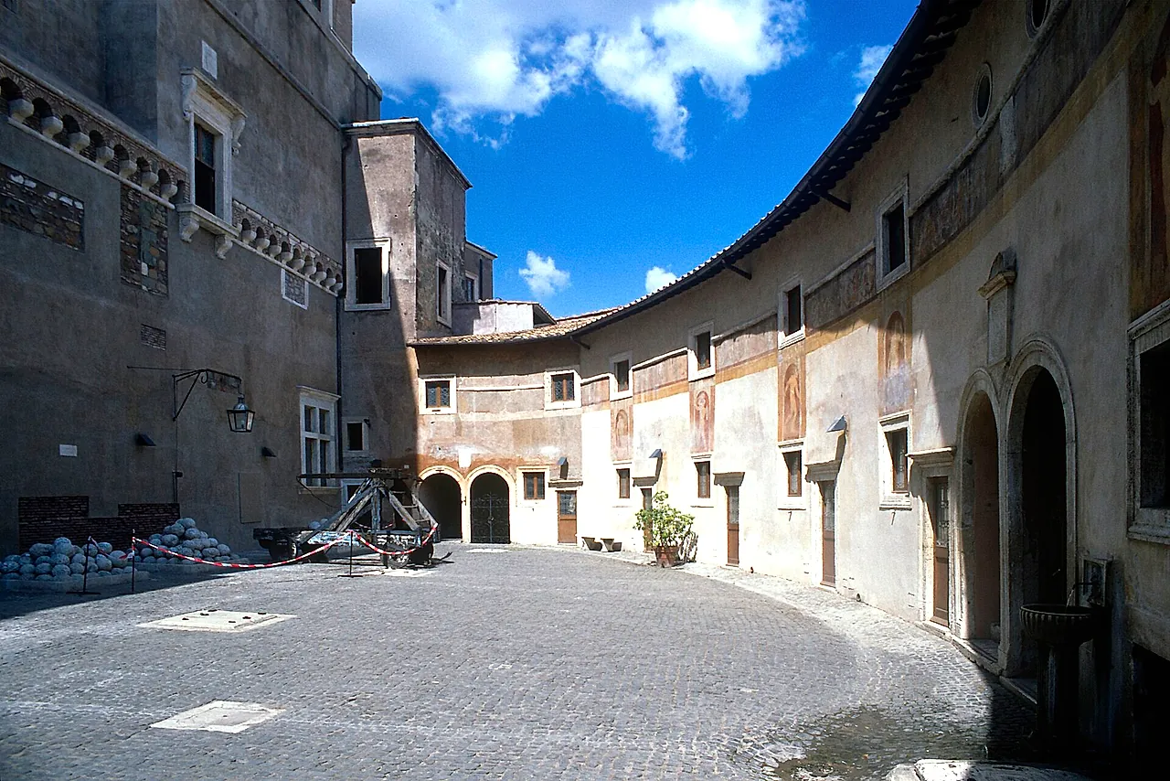 The curved Courtyard of Alexander VI inside the castle, with faded frescoes on the walls, a pile of stone cannonballs and a wooden catapult, under a blue sky with a few clouds