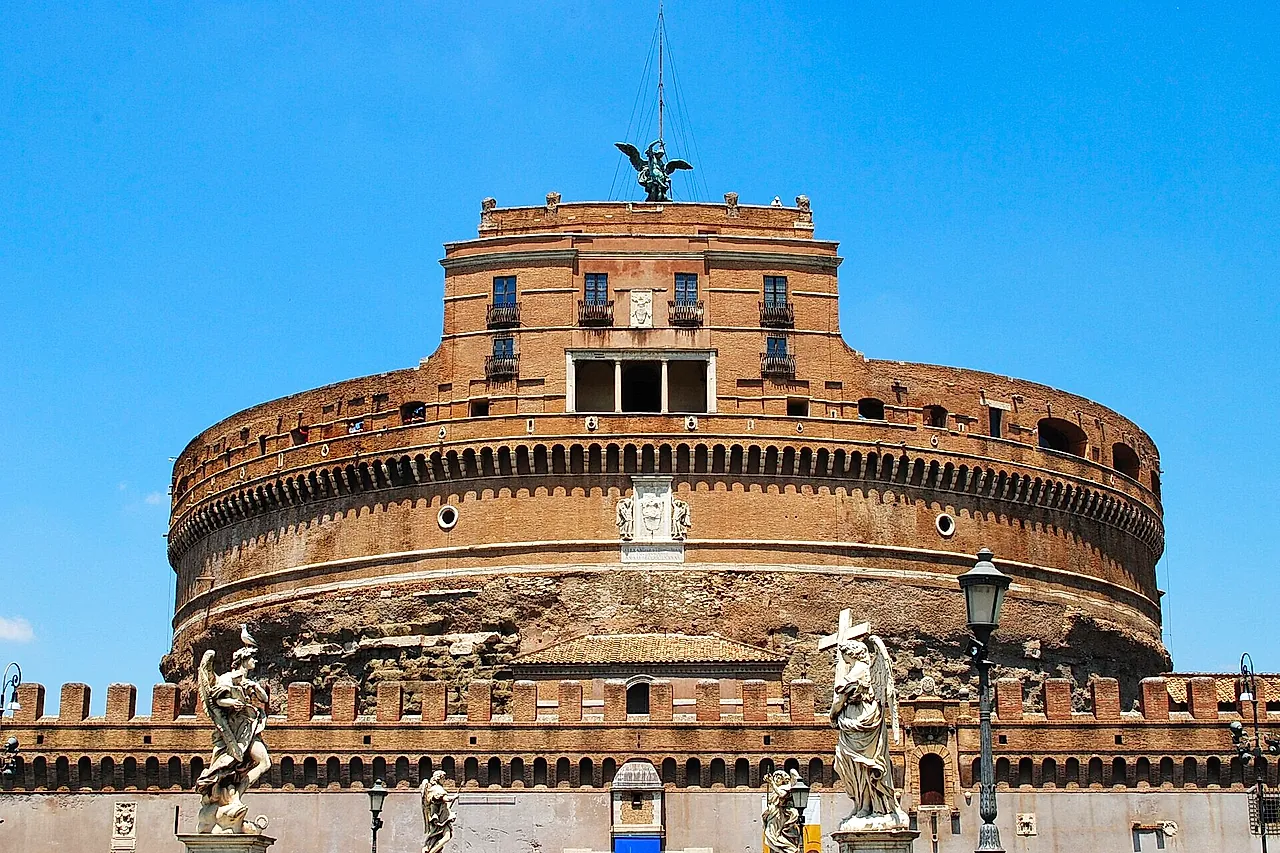 Castel Sant'Angelo seen from the Ponte Sant'Angelo against a bright blue sky: the round brick drum of the old mausoleum, the square upper block with the bronze angel on top and the marble angel statues of the bridge in the foreground