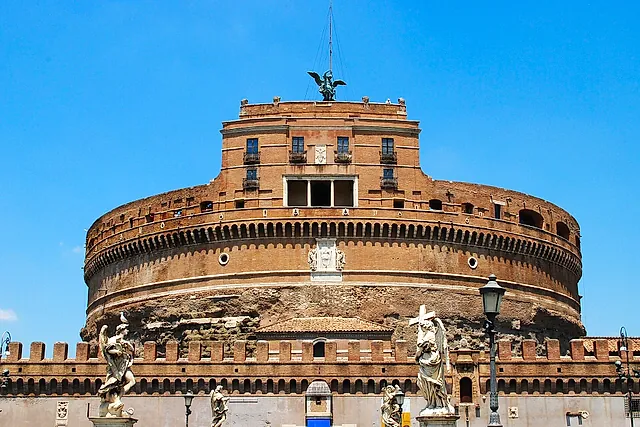 Castel Sant'Angelo seen from the Ponte Sant'Angelo against a bright blue sky: the round brick drum of the old mausoleum, the square upper block with the bronze angel on top and the marble angel statues of the bridge in the foreground