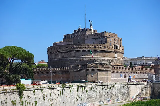 Castel Sant'Angelo seen from the Tiber embankment on a sunny day: the massive round drum and the battlemented bastions above the river wall, with a stone pine on the left