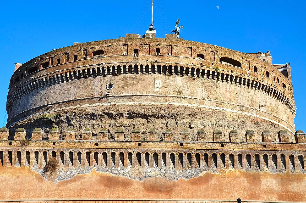 Close view of the huge round drum of Castel Sant'Angelo, Hadrian's old mausoleum, with its rows of small arches and the battlemented outer wall in front, under a blue sky