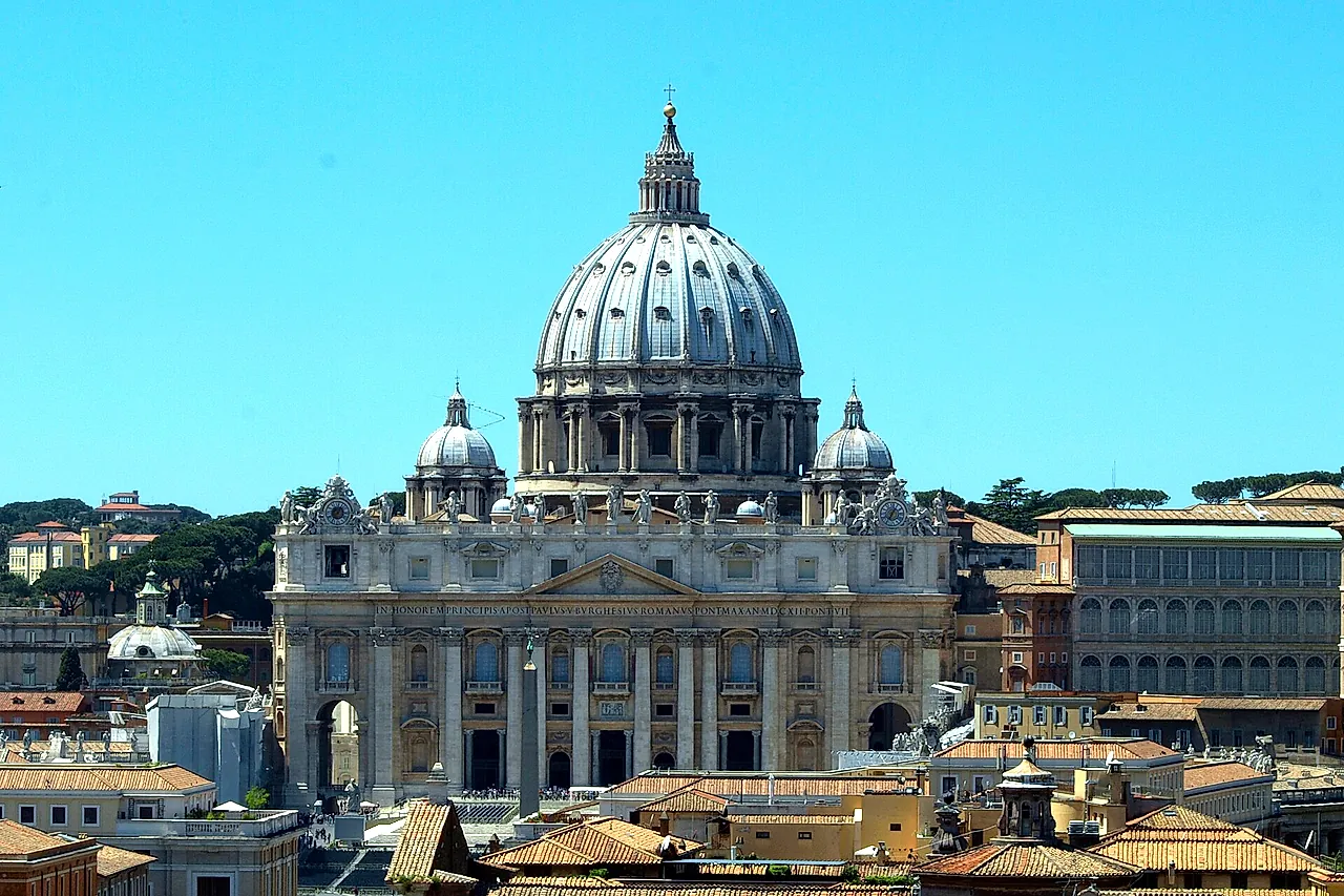 St. Peter's Basilica and its dome seen from Castel Sant'Angelo over the terracotta rooftops of the Borgo district, under a clear blue sky
