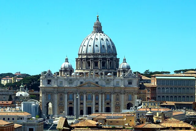 St. Peter's Basilica and its dome seen from Castel Sant'Angelo over the terracotta rooftops of the Borgo district, under a clear blue sky