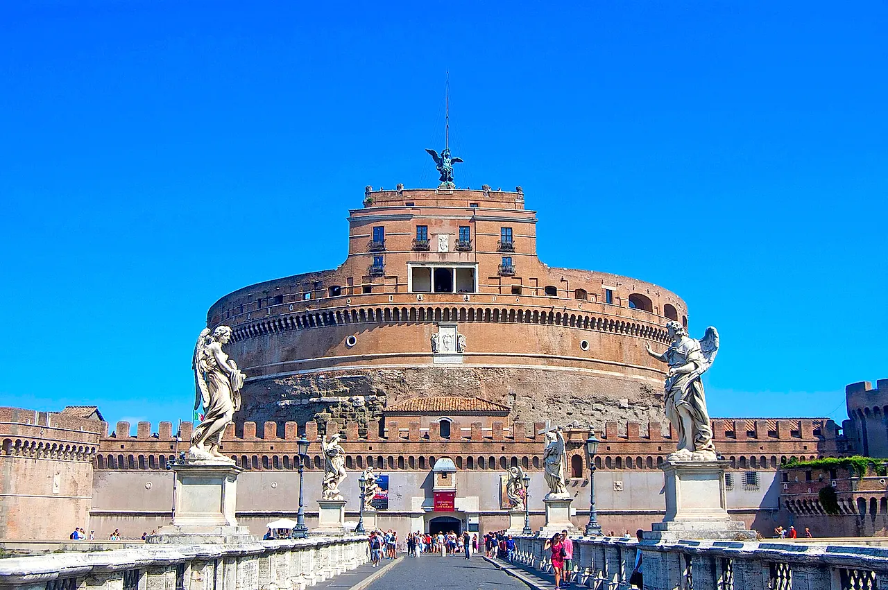 The Ponte Sant'Angelo leading to the castle, with two marble angel statues on their pedestals in the foreground, visitors walking on the bridge and a deep blue sky