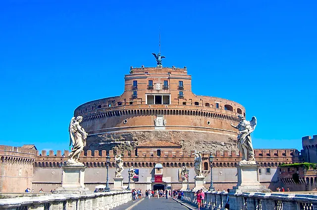 The Ponte Sant'Angelo leading to the castle, with two marble angel statues on their pedestals in the foreground, visitors walking on the bridge and a deep blue sky