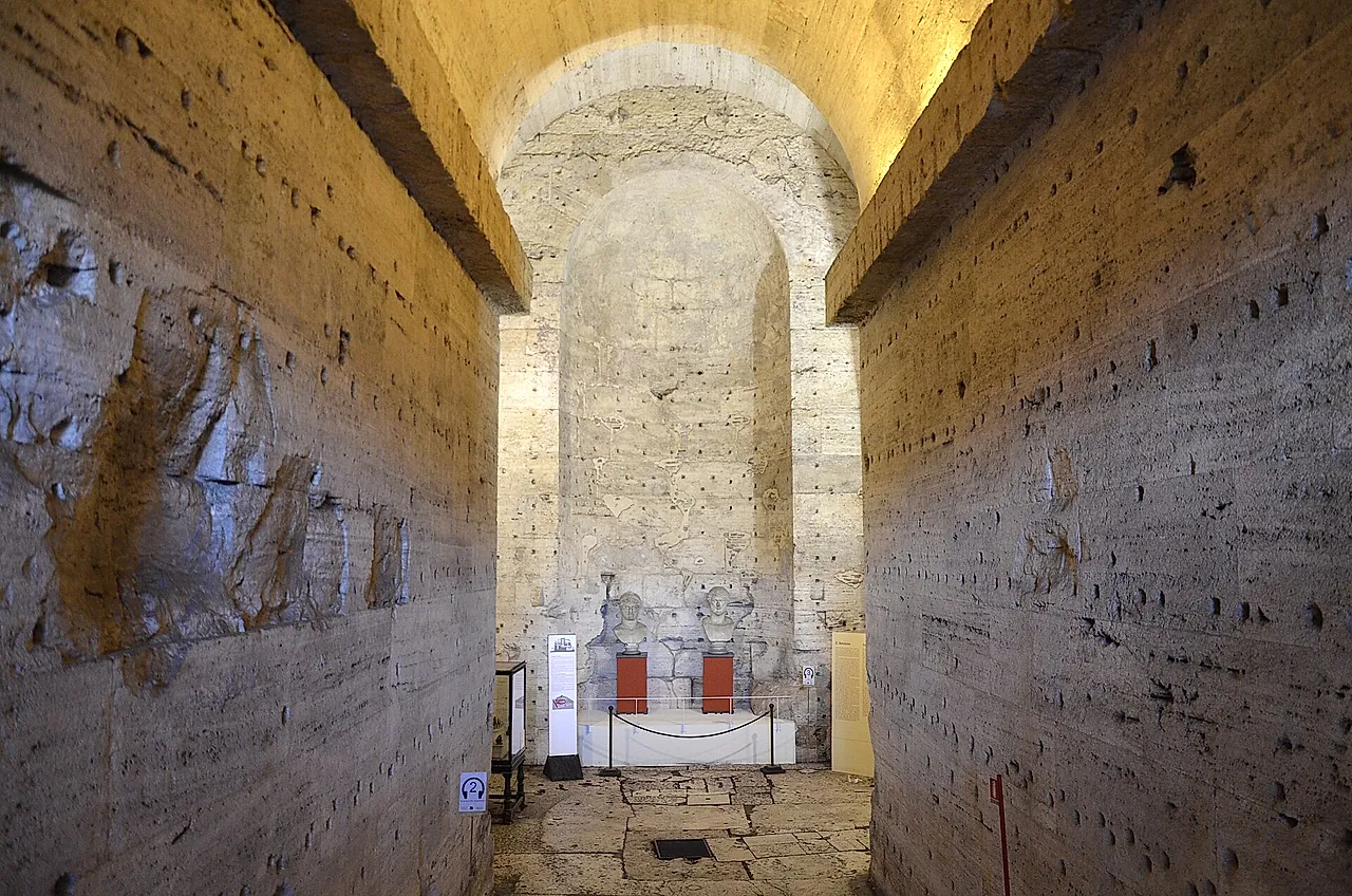 A long straight corridor of rough stone and brick inside the old mausoleum, rising toward a lit arched niche at the far end where a small display stands behind a rope