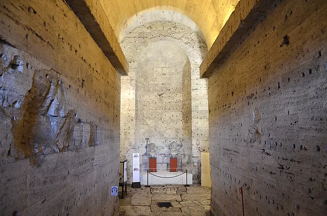 A long straight corridor of rough stone and brick inside the old mausoleum, rising toward a lit arched niche at the far end where a small display stands behind a rope