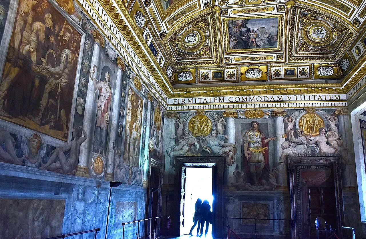 The Sala Paolina, the Renaissance hall of the castle, with a gilded coffered ceiling, frescoed walls and a bright open doorway where two visitors stand as silhouettes