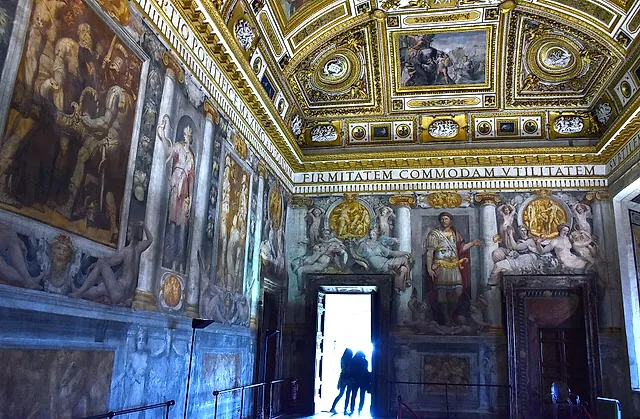 The Sala Paolina, the Renaissance hall of the castle, with a gilded coffered ceiling, frescoed walls and a bright open doorway where two visitors stand as silhouettes