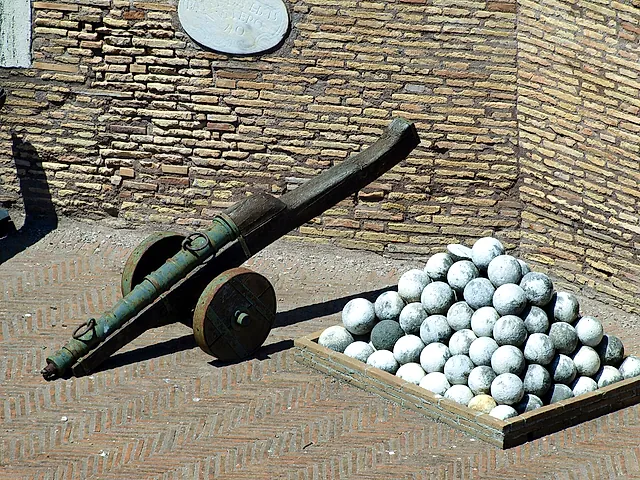 An old cannon with a green bronze barrel on a wooden carriage next to a pile of round stone cannonballs in a wooden tray, on a herringbone brick floor in front of a brick wall