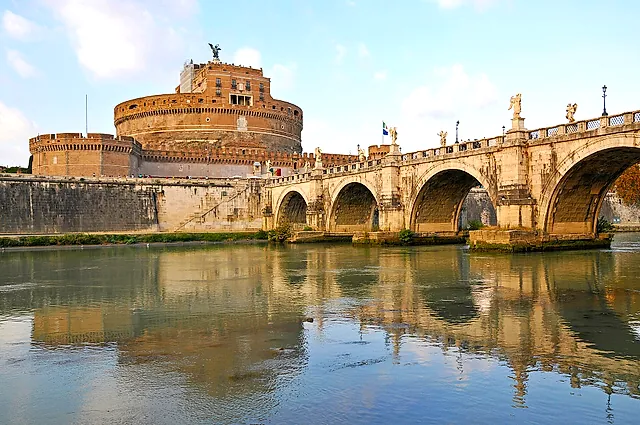 Castel Sant'Angelo and the Ponte Sant'Angelo seen from the Tiber embankment, with the arches and the castle reflected in the calm water under a sky of light clouds