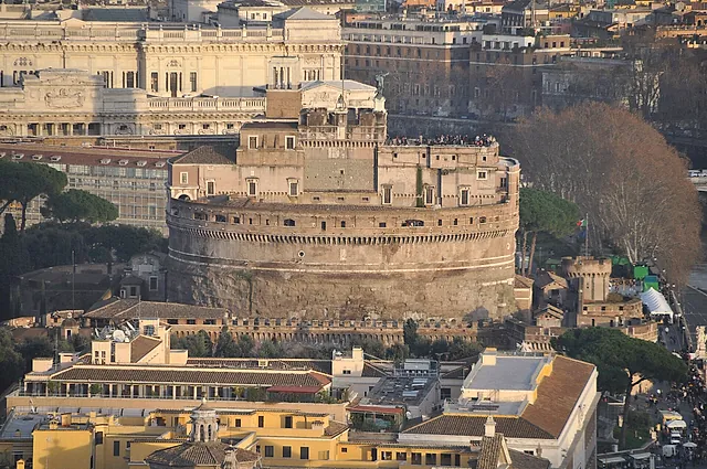 High-angle view of Castel Sant'Angelo in warm light, with the round drum, the upper terrace where tiny visitors stand and the rooftops of Rome and a large ornate palace behind it