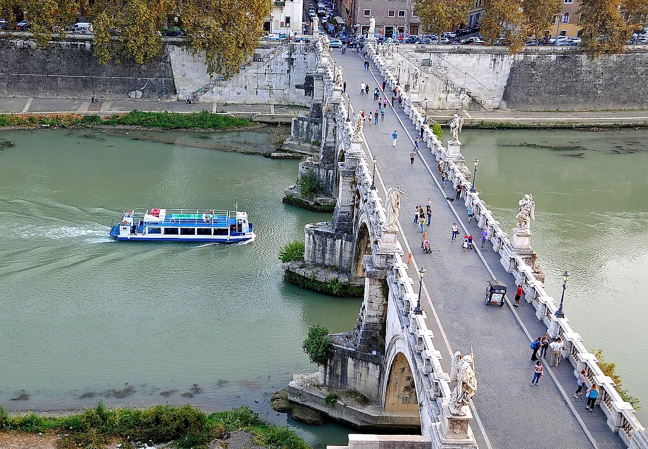 View from the castle looking down on the Ponte Sant'Angelo and the Tiber, with a tour boat passing and people walking among the angel statues of the bridge