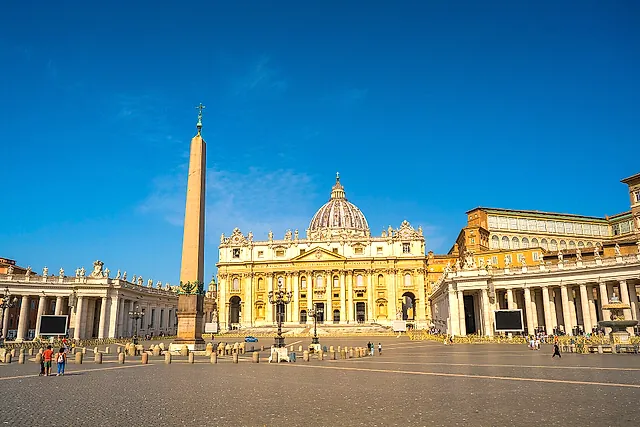 St. Peter's Square in warm light, with the Egyptian obelisk on the left, the colonnades on both sides and the Basilica with its dome in the background under a blue sky