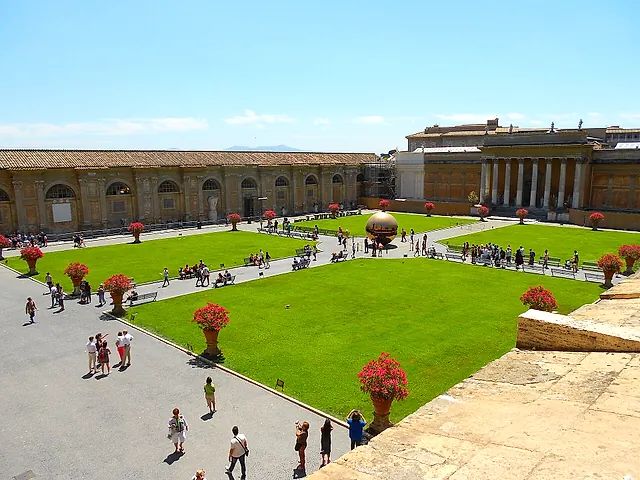 The Cortile della Pigna in the Vatican Museums seen from above: green lawns with red flowering pots, gravel paths with a few visitors, the long gallery wall and a large golden sphere sculpture in the distance